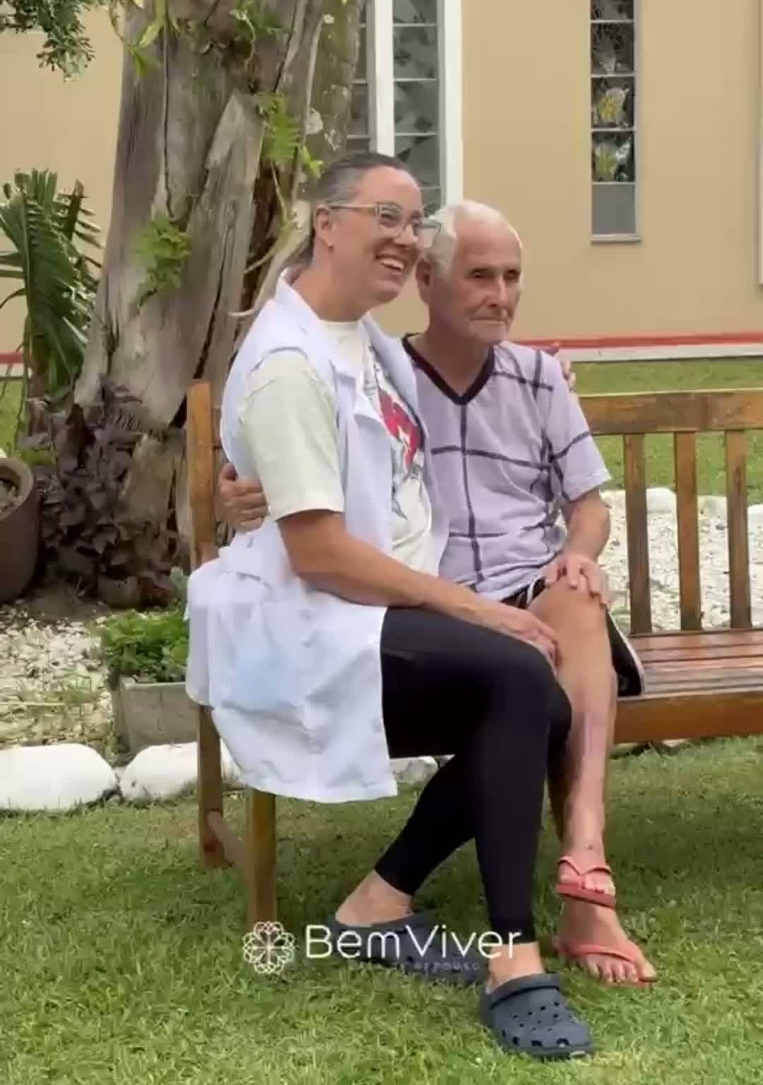 Cuidadora sorrindo ao lado de idoso sentado em banco no jardim da casa